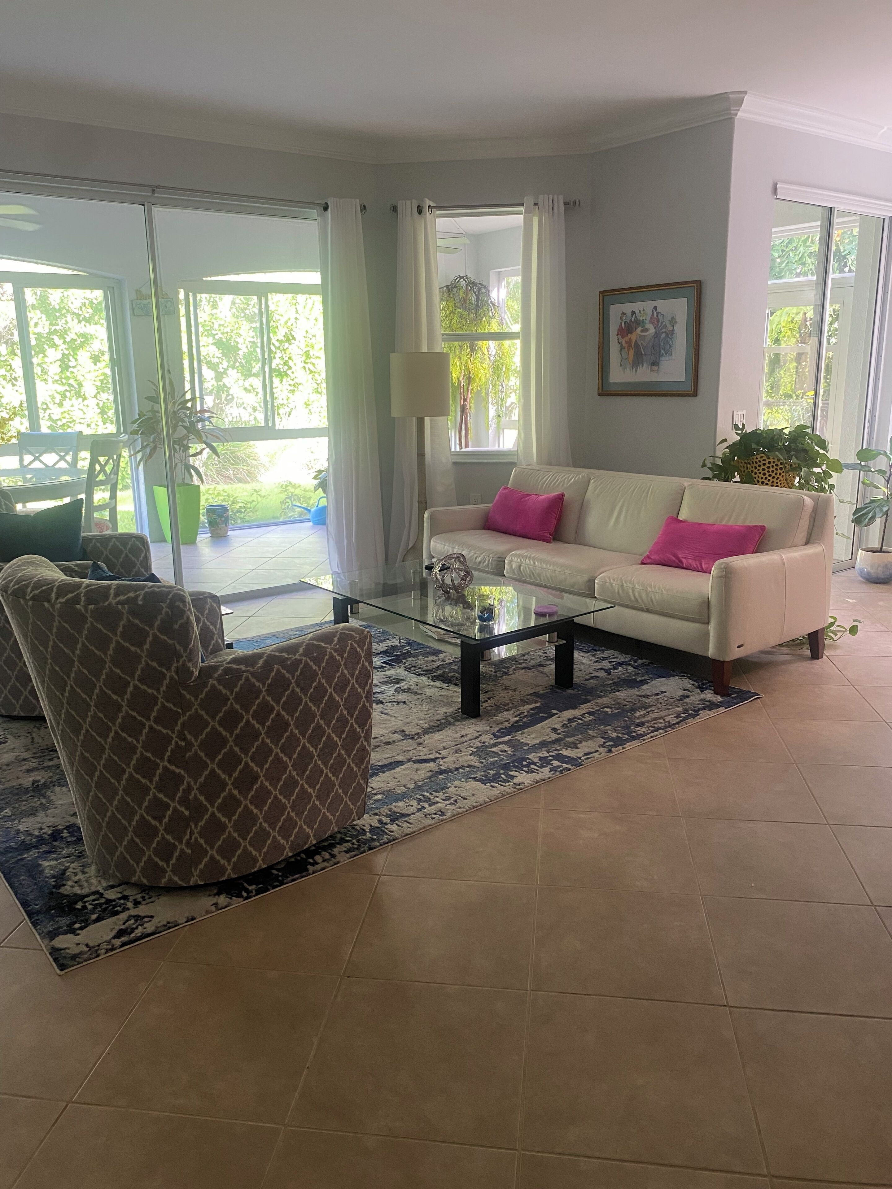 A living room with a beige sofa, a patterned armchair, a glass coffee table, and a blue and white rug. There is a sliding glass door leading to an outdoor area with a chair and a potted plant.