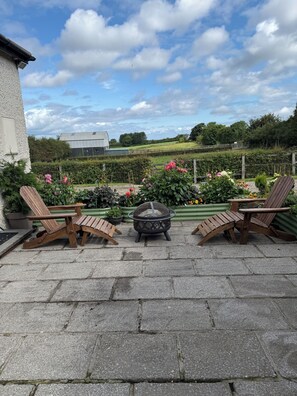 Eine Terrasse mit Holzstühlen, einer Feuerstelle und Blick auf ein Bauernhofgebiet und Hügel.