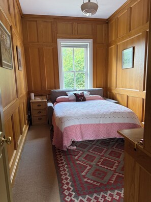 A bedroom with wooden paneling, a bed with a patterned bedspread, a window with a view of greenery, a bedside table, and a nightstand.