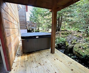A wooden deck with a hot tub, surrounded by a natural landscape with trees and a stream. - Keystone Lodge A | Ski-In/Out, Hot Tub! (Snowshoe)