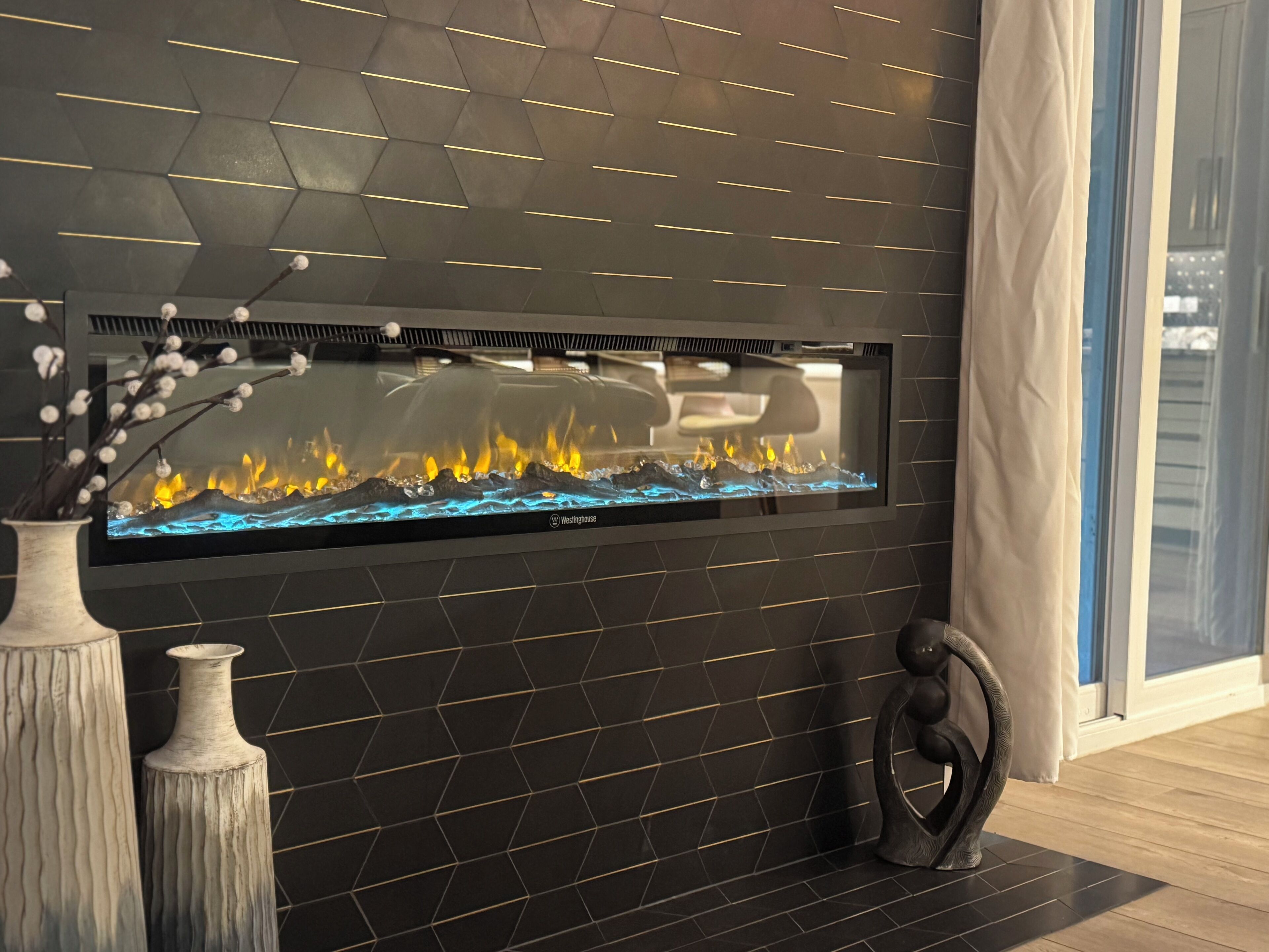 A modern fireplace with a glass front, set in a black tiled wall with geometric patterns.