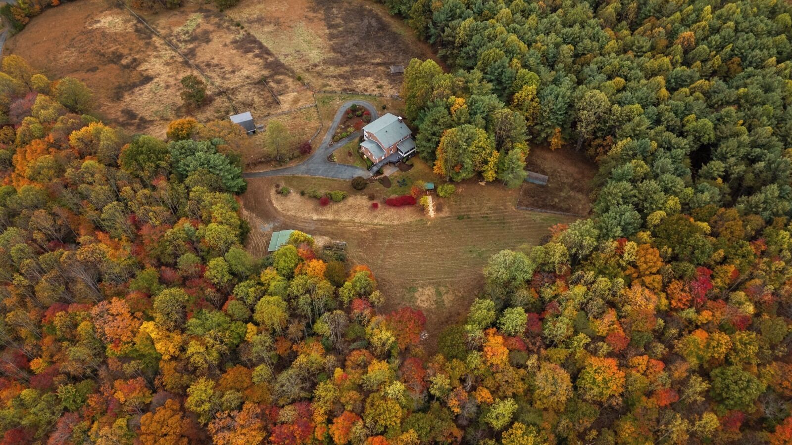 An aerial view of a rural property with a house surrounded by dense forest, a driveway, and a small fenced area.