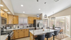 Una cocina moderna con armarios de madera, electrodomésticos de acero inoxidable, una isla central con taburetes y amplias ventanas con persianas.