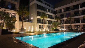 Een modern hotel met een zwembad, palmbomen en verlichte balkons.