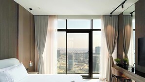 Ein modernes Hotelzimmer mit einem großen Bett, einem Schreibtisch mit Fernseher und Blick auf eine Skyline durch raumhohe Fenster.