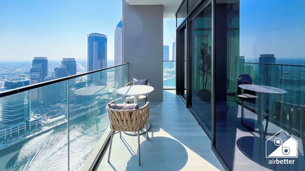 Ein moderner Balkon mit Glasgeländer, einem Korbstuhl, einem kleinen runden Tisch und Blick auf eine Stadtlandschaft.