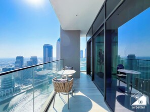 A modern balcony with a glass railing, a wicker chair, a small round table, and a view of a cityscape. - Luxurious 3BR Canal View Apartment Pool + Gym (Dubai)