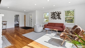 Un salon spacieux doté d’un parquet, d’un canapé rouge, d’un fauteuil blanc et d’une table basse. Deux fenêtres laissent entrer la lumière naturelle, et un grand tableau abstrait orne le mur.
