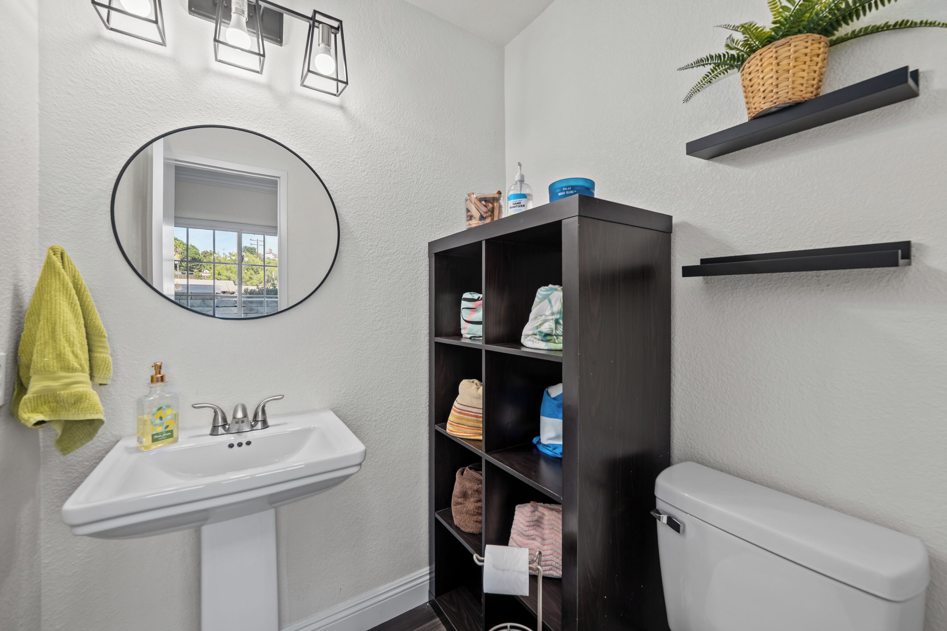 A bathroom with a white sink, a mirror, a toilet, and a dark wooden shelf with toiletries.