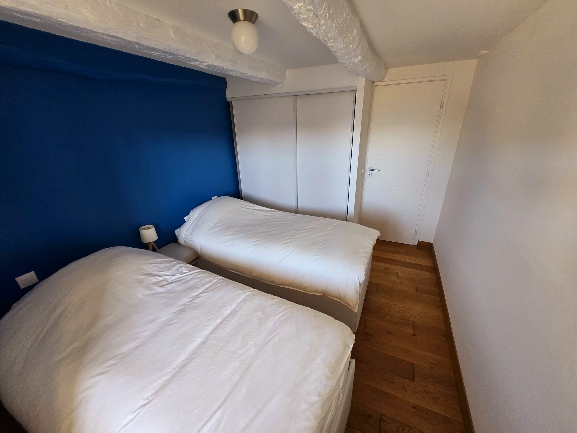 A bedroom with two beds, a blue accent wall, a wooden floor, and a white door.