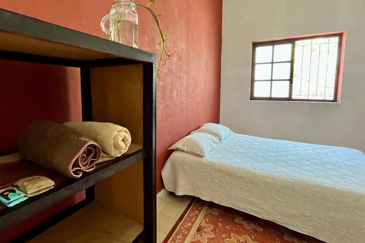 A room with a bed, a shelf with a rolled towel, a window with bars, and a plant in a glass jar.