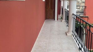 Un corridoio con pavimento piastrellato, una porta in legno e un balcone con ringhiera nera.