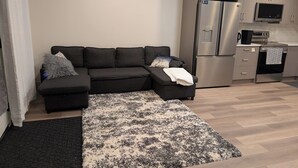 A modern living room with a sectional sofa, a patterned rug, and a kitchen area with stainless steel appliances.