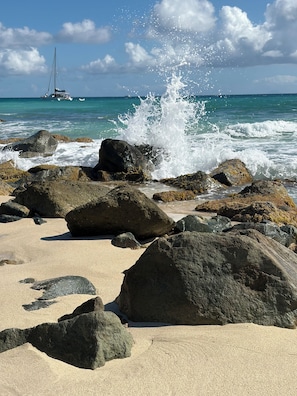 A beach with large rocks, waves crashing, and a sailboat in the distance.