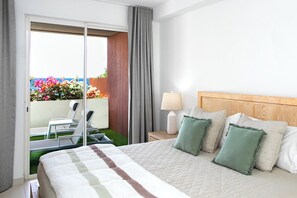 A hotel room with a bed, pillows, a nightstand, a lamp, a chair, and a view of a balcony with flowers.