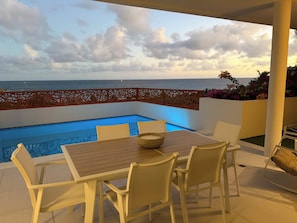 A poolside area with a table and chairs, a view of the ocean, and a decorative fence.