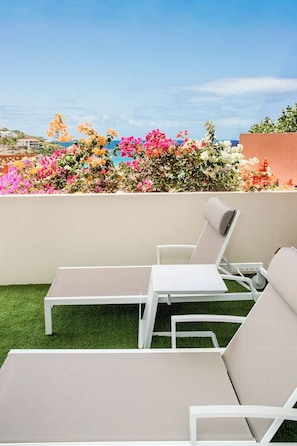 A rooftop terrace with two white lounge chairs, a small table, and a view of colorful flowers and a clear sky.