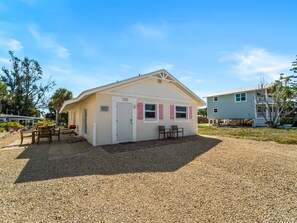 A small, single-story building with a white facade and a red door, labeled '1125', with a gravel driveway and outdoor seating area. - Seahorse Cottage 2 - 2/2 with Private Beach (Englewood)