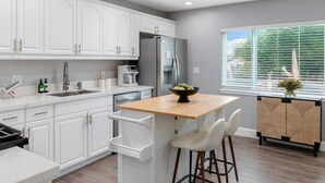 A modern kitchen with white cabinetry, a stainless steel refrigerator, a coffee maker, and a window with blinds. - Bella| Pool/Spa|Downtown|Freeways|Beaches (San Diego)