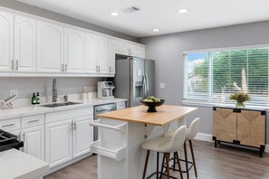 A modern kitchen with white cabinetry, a stainless steel refrigerator, a coffee maker, and a window with blinds. - Bella| Pool/Spa|Downtown|Freeways|Beaches (San Diego)