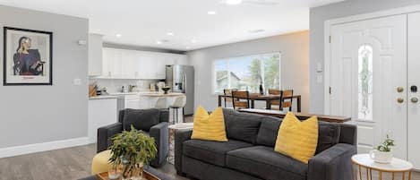 A modern living room with a gray sofa, a dining table, and a kitchen in the background.