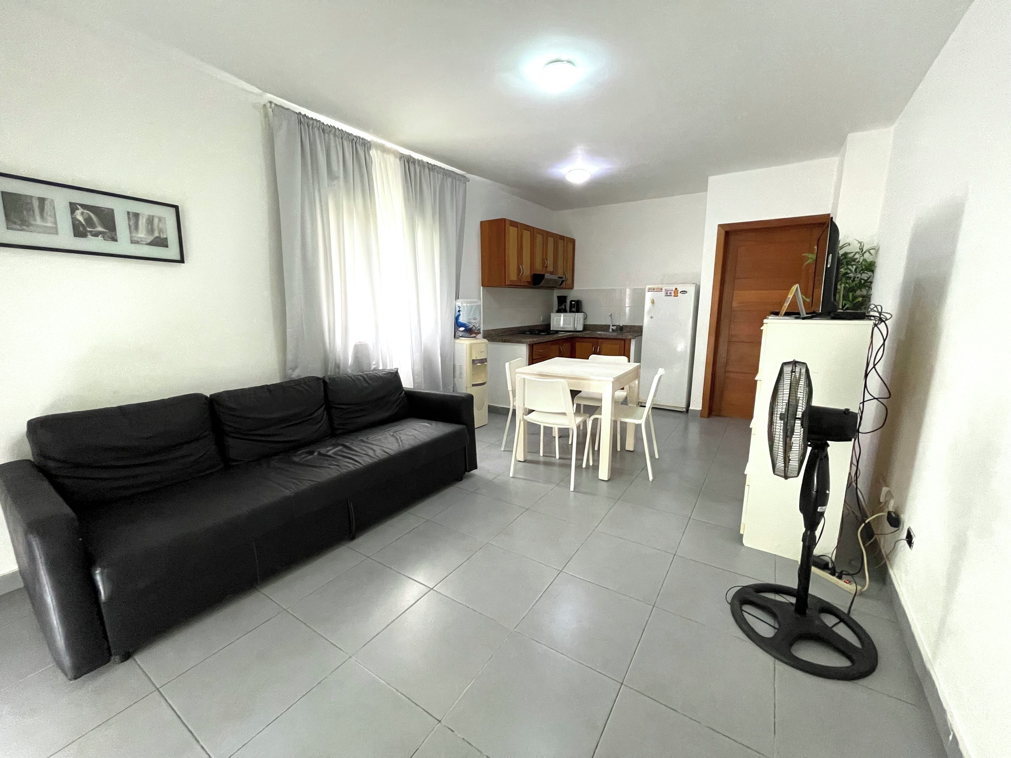 A living room with a black sofa, a dining table with chairs, a kitchen area with cabinets and appliances, and a standing fan.
