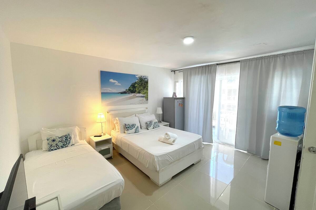 A hotel room with two beds, a desk, a lamp, a water dispenser, and a painting of a beach scene.