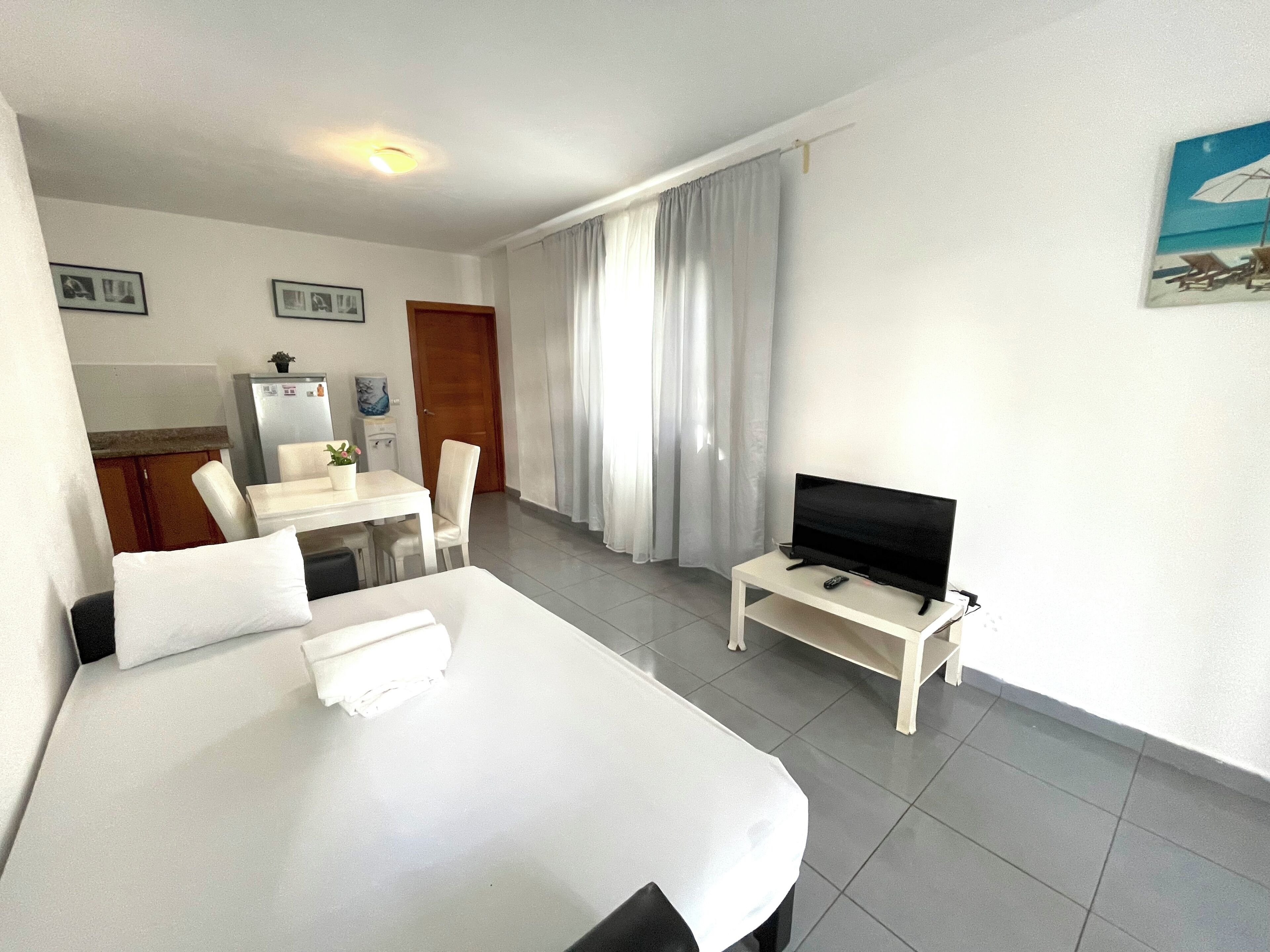 A hotel room with a bed, a TV on a small table, a desk with a chair, and a kitchenette in the background.
