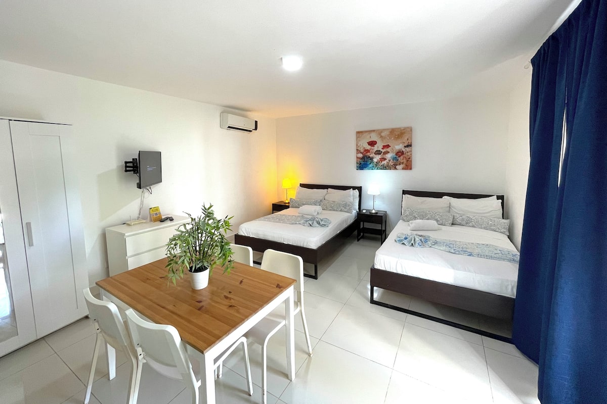 A hotel room with two beds, a wooden table with chairs, a desk, a TV, and a painting on the wall.