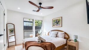 Un dormitorio con una cama de madera, un ventilador de techo, una ventana con vista a una cerca y un cuadro enmarcado en la pared.