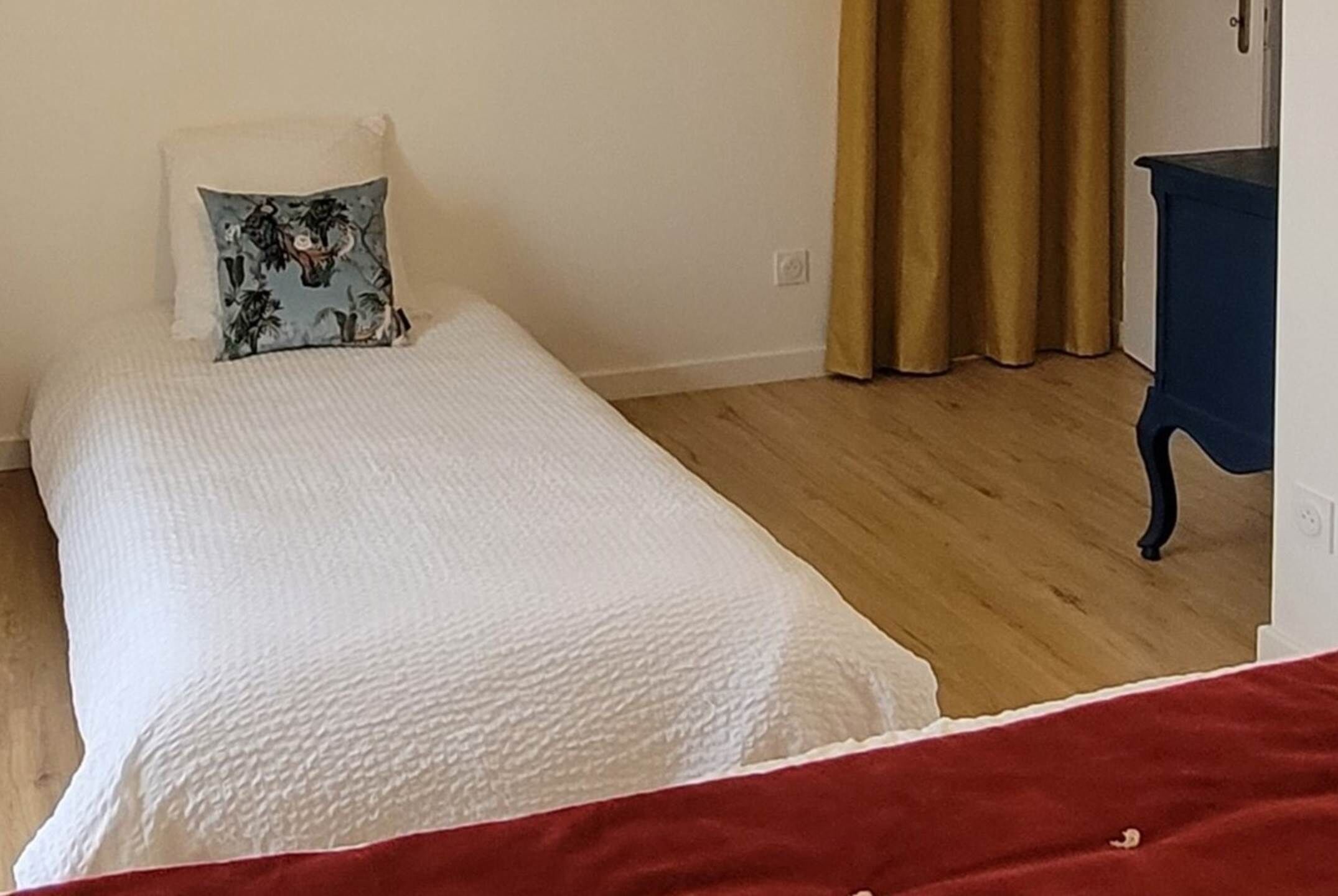 Un letto singolo con copriletto bianco e cuscino decorativo, pavimento in legno, una cassettiera blu e una tenda beige.