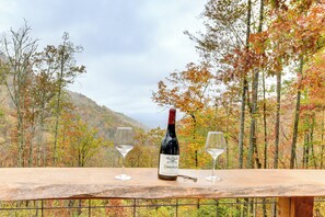 A bottle of wine and two glasses on a wooden table with a scenic view of autumn trees and hills in the background.