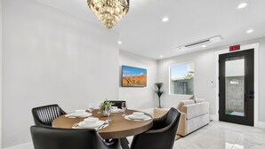 Et moderne spiseområde med et rundt bord dækket til fire personer, sorte læderstole og en lysekrone. Der er en sofa og et vindue med udsigt over et ørkenlandskab.