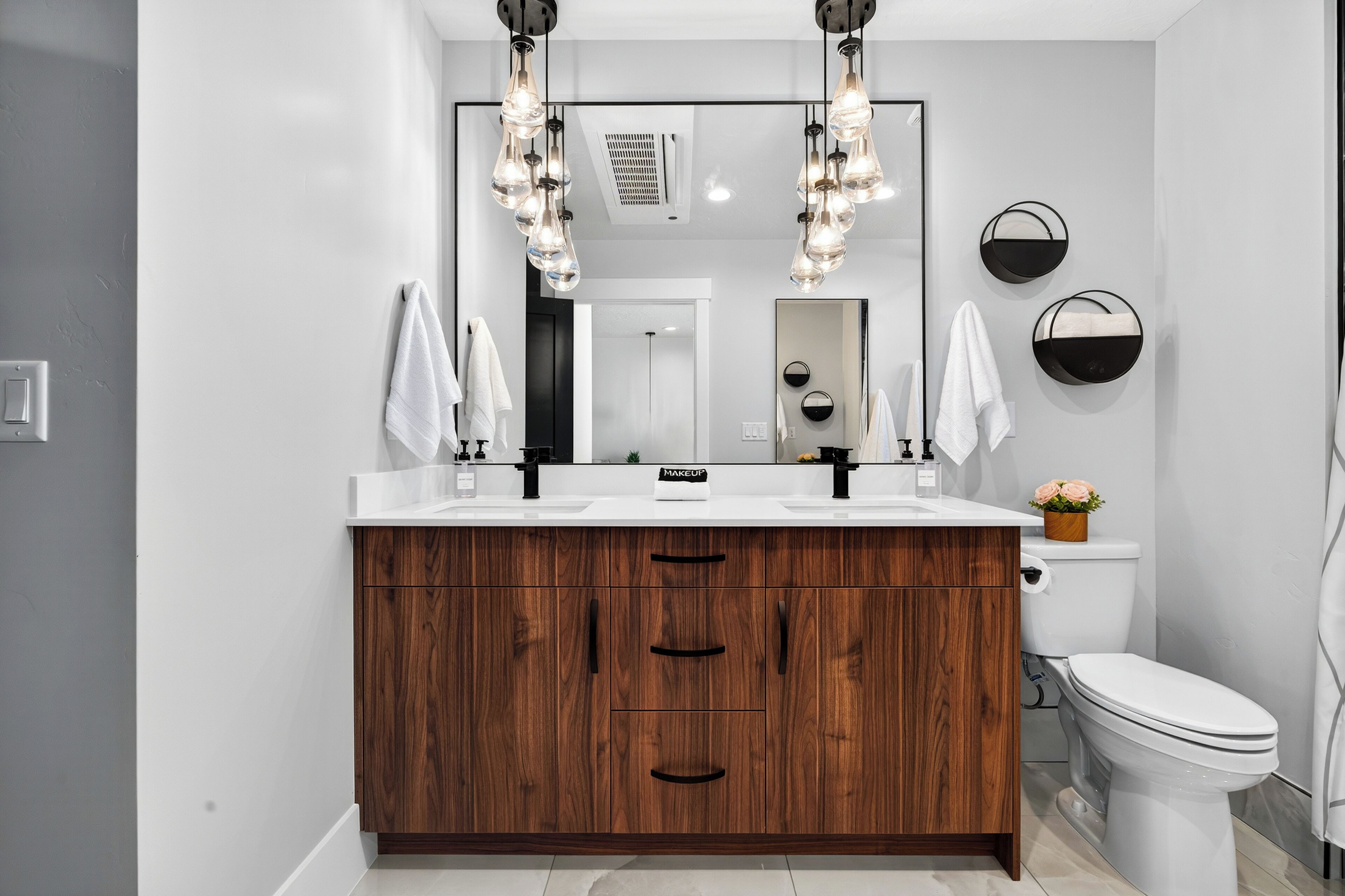 A modern bathroom with a wooden vanity, two sinks, a toilet, and a large mirror with light fixtures.
