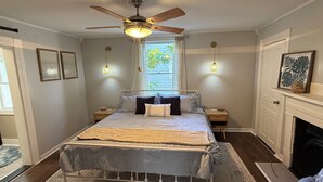Un dormitorio con cama, mesitas de noche, ventilador de techo, ventana con cortinas, chimenea y cuadros en la pared.