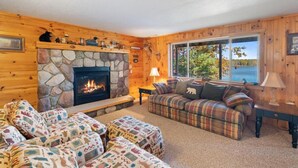 A cozy cabin interior with a stone fireplace, plaid sofa, and a view of a lake. - 4BR Lakefront Home | Kayaks, Fire Pit & Views (Hawks)