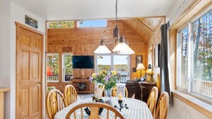 A wooden cabin interior with a dining table, chairs, a TV, and a window with a view of trees. - 4BR Lakefront Home | Kayaks, Fire Pit & Views (Hawks)