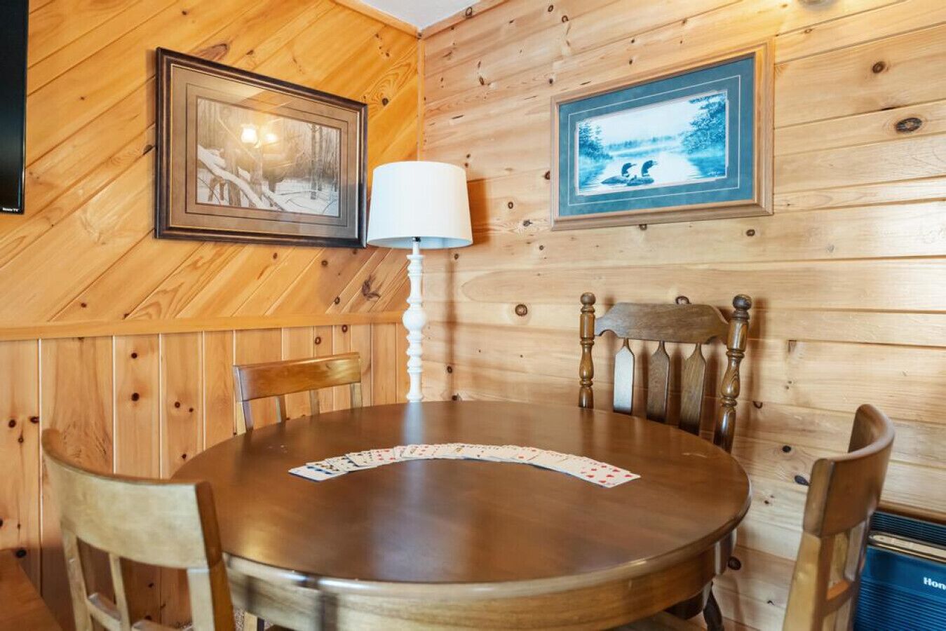 Un interior de madera con una mesa redonda, sillas, una lámpara y cuadros en las paredes.