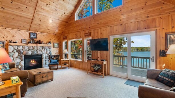 A cozy cabin interior with a stone fireplace, a television, and a large window overlooking a body of water. - 4BR Lakefront Home | Kayaks, Fire Pit & Views (Hawks)