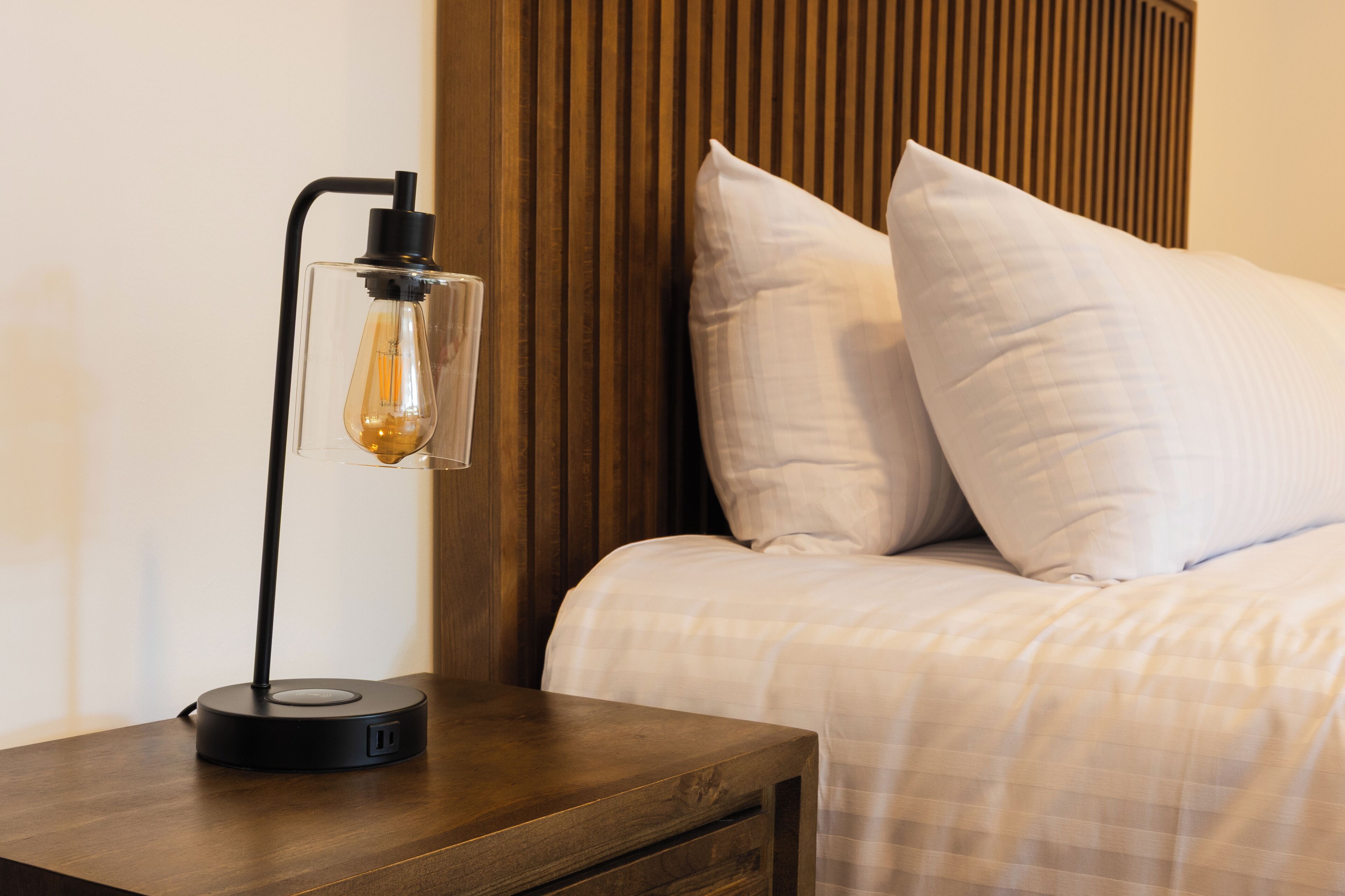 Ein ordentlich bezogenes Bett mit Holz-Kopfteil, ein Nachttisch mit Lampe sowie eine Wand mit vertikalen Holzbalken.