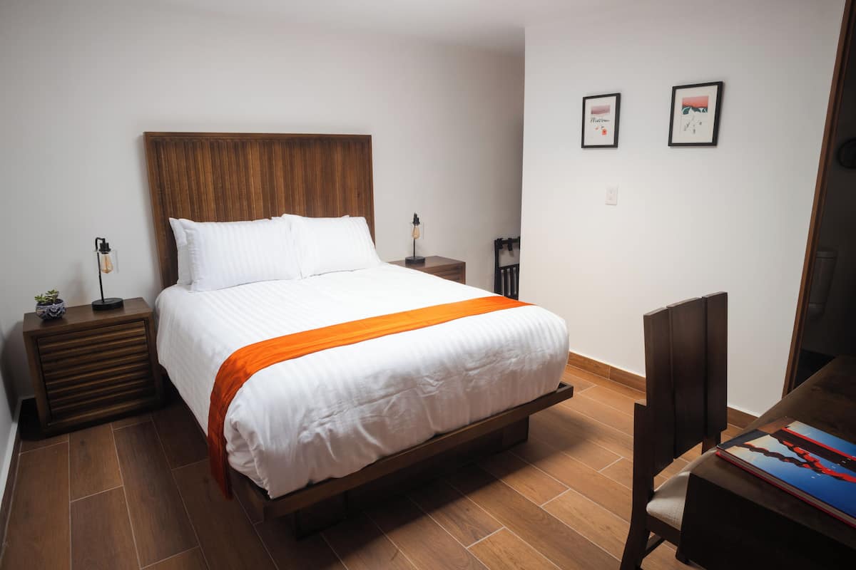 Una cama bien hecha con cabecera de madera, una mesita de noche con una lámpara y una silla con un libro.