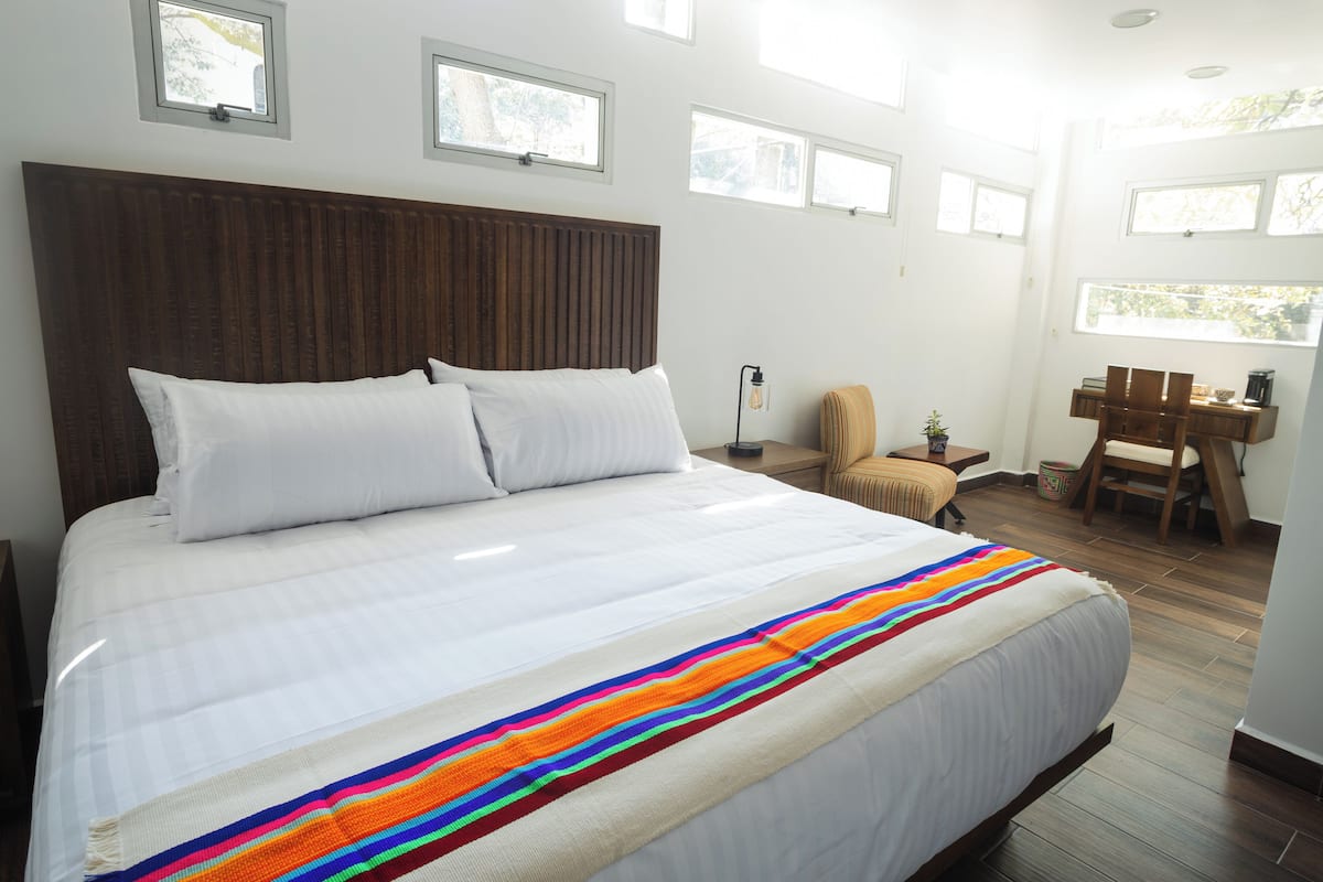 Una cama grande con cabecera de madera, ropa de cama blanca y una manta a rayas coloridas. Varias ventanas proporcionan luz natural, una mesita con una silla y un escritorio con una silla y una planta.