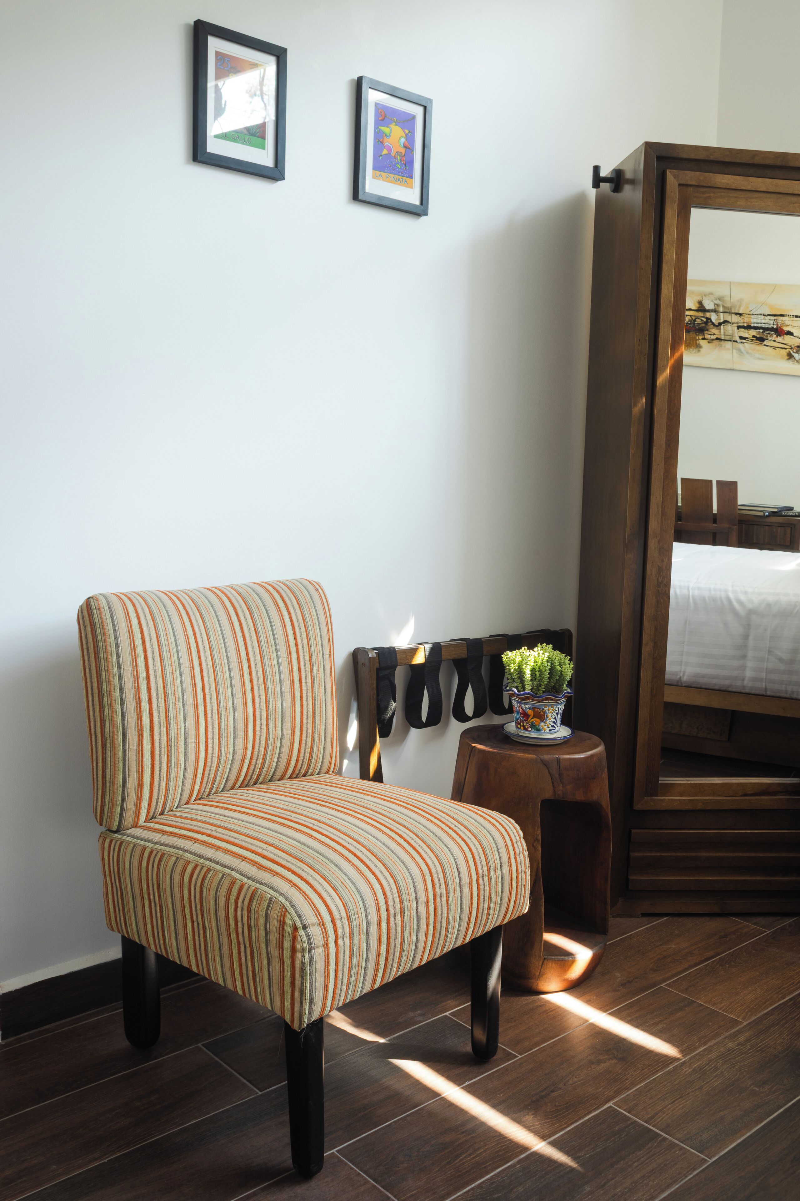 A striped armchair, a small wooden stool with a plant, and a bed with a wooden headboard in a room with dark wooden flooring and white walls.