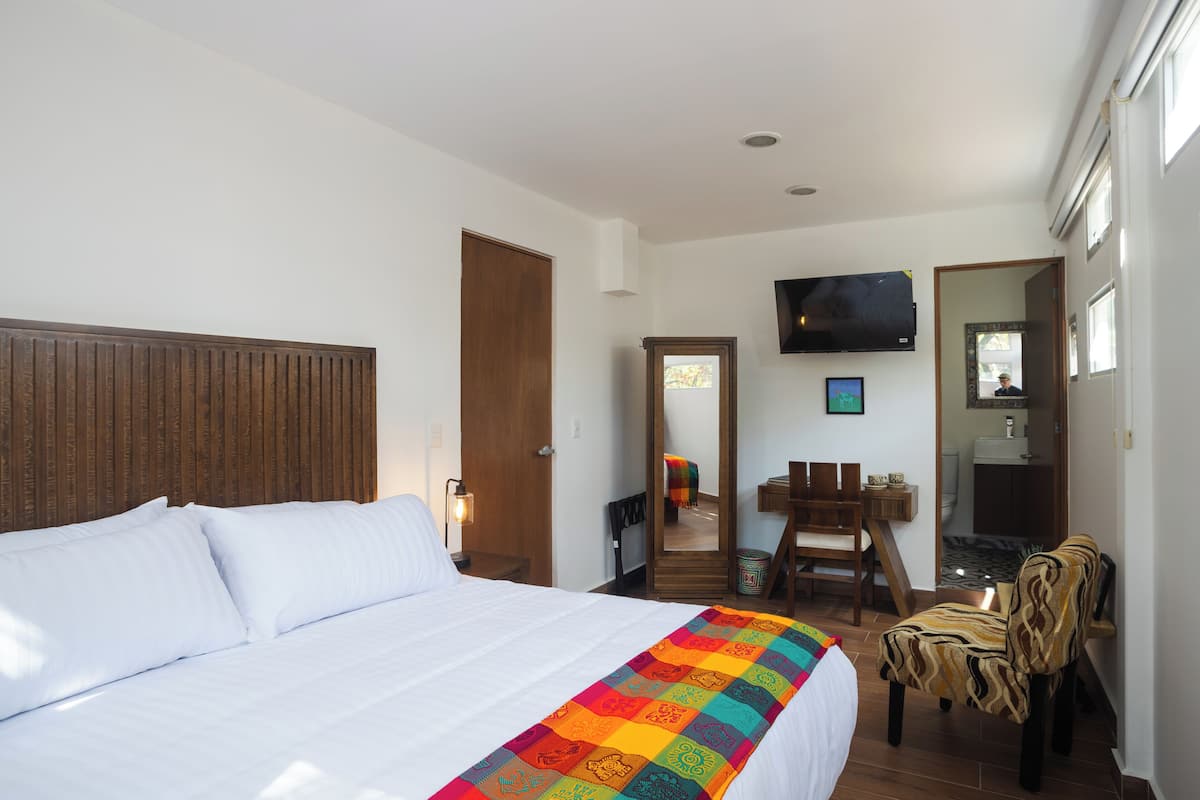 Habitación de hotel con una cama grande, cabecera de madera, televisión montada en la pared, un espejo y una pequeña mesa con sillas.