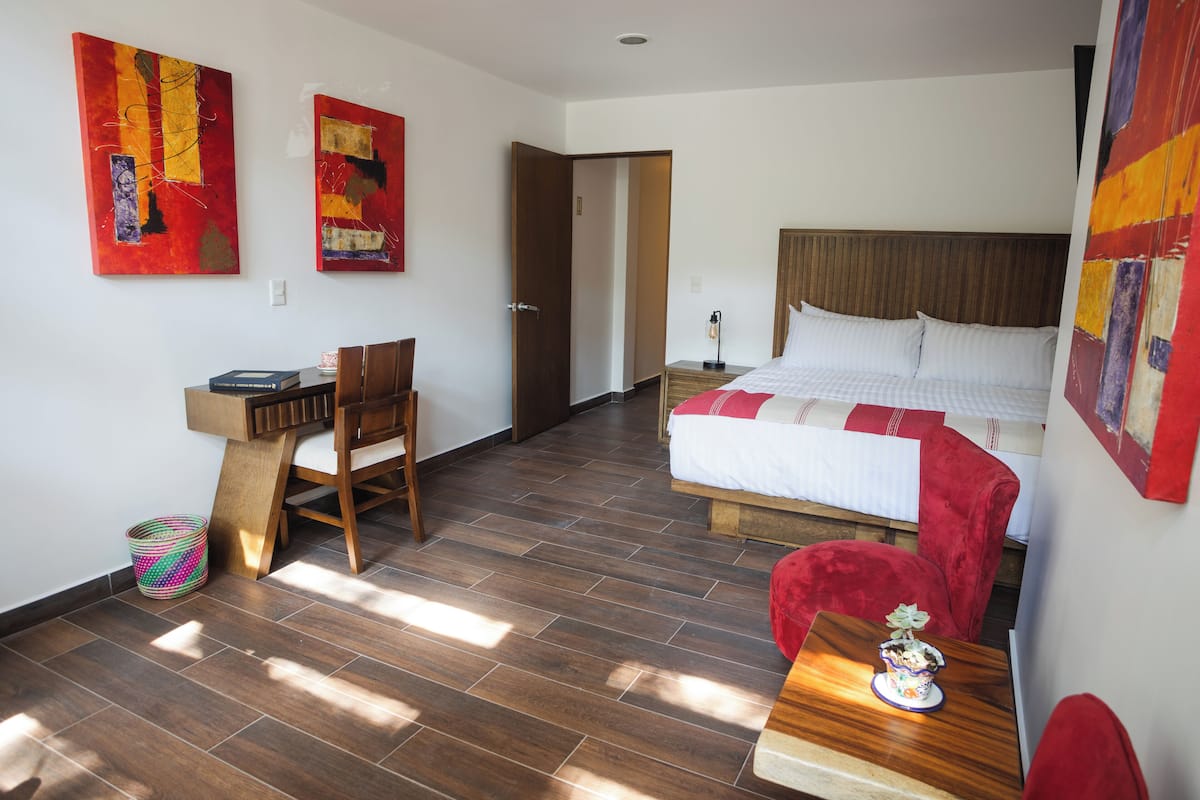 Habitación de hotel con piso de madera, cama con ropa blanca, escritorio con silla y dos cuadros abstractos en las paredes.