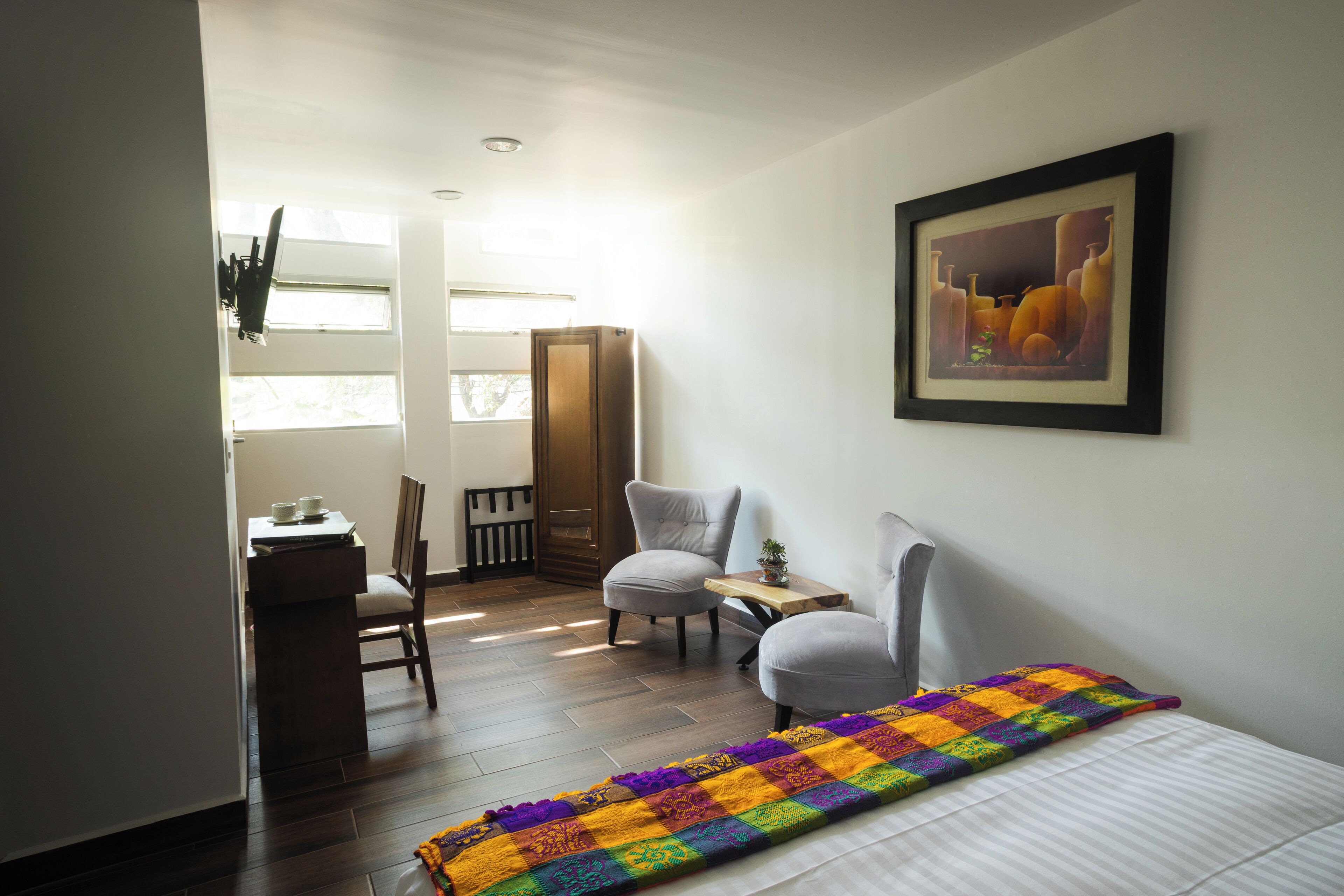 Une chambre d’hôtel avec un lit, deux chaises, un bureau, une petite table avec une plante et un tableau encadré au mur.