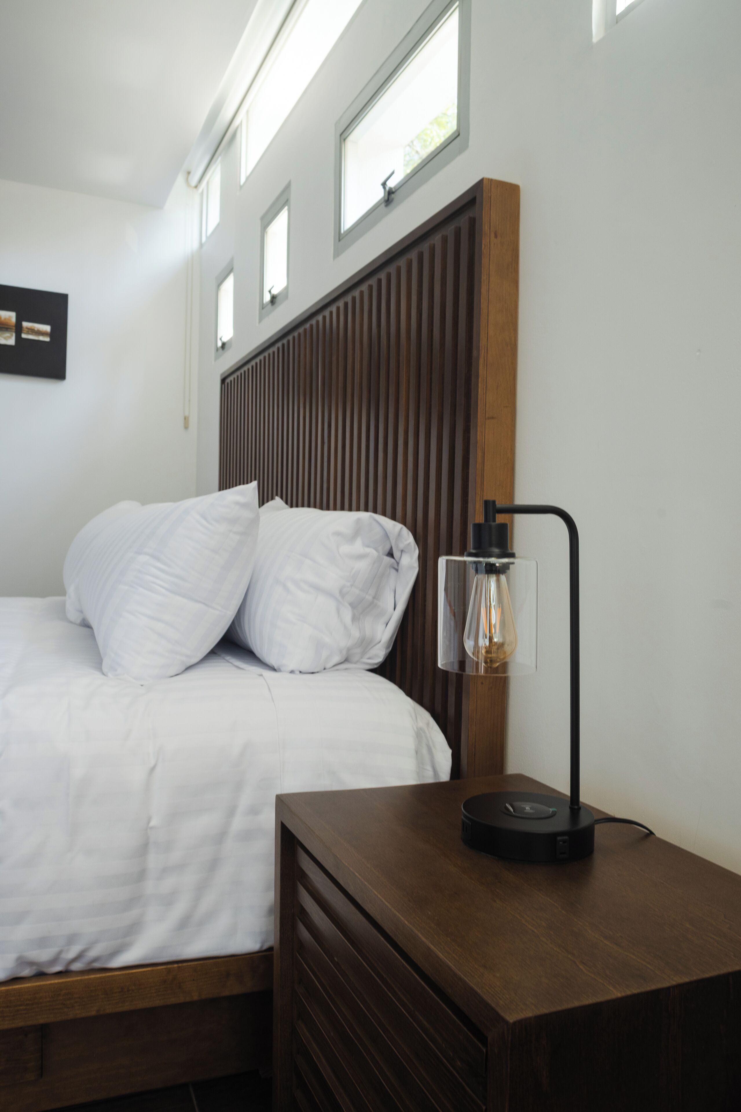 A neatly made bed with white linens and pillows, a wooden headboard, a bedside table with a lamp, and a small window above the bed.