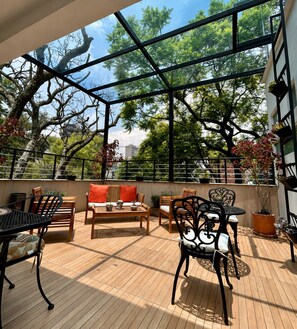 A spacious outdoor patio with wooden flooring, metal and wooden furniture, and large windows offering views of trees and buildings. - La Casa del Mural (Mexico City)