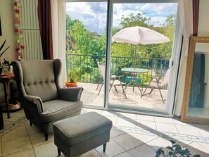 Ein Wohnzimmer mit grauem Sessel, einem kleinen Hocker und einer Schiebetür zu einer Außenpatio mit Tisch und Stühlen sowie einem großen Sonnenschirm.