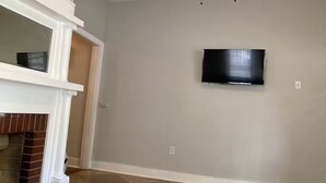 A room with a flat-screen TV mounted on the wall, a white mantelpiece, and a checkered floor. - Cozy Apartment in the heart of town. Monthly min. (Bowling Green)